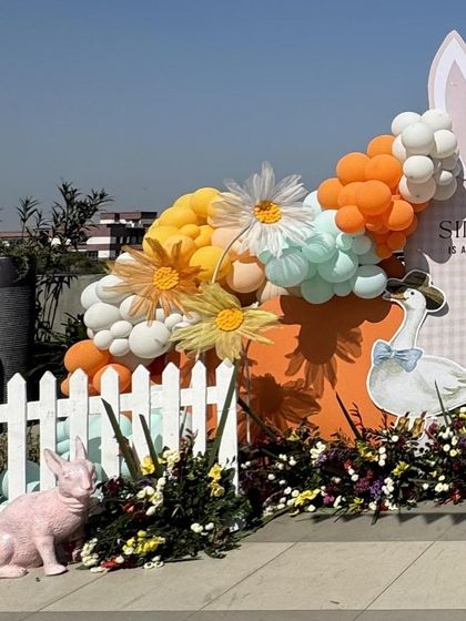 A close-up of the bunny garden backdrop, showing a white picket fence, colorful florals, and a playful arrangement of orange, yellow, and mint green balloons.