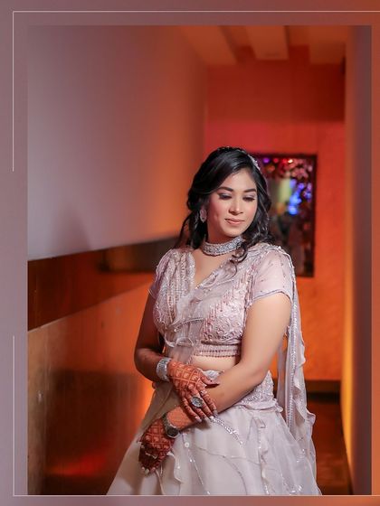 A solo portrait of the bride at her Roka ceremony. She looks down gracefully, showcasing her intricate lehenga, henna-adorned hands, and sparkling jewelry in a softly lit hallway.