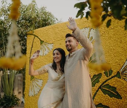 The couple waving against a vibrant marigold backdrop, their coordinated neutral outfits creating a calm and elegant picture.