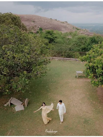 A high-angle shot of a couple walking through a grassy area near the Melukote hills, capturing their movement and joy.