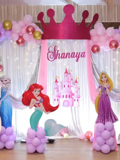A Disney Princess theme party fit for royalty. This backdrop features cutouts of Ariel, Rapunzel, and Aurora, a castle, and a large glittery crown, all framed by pink, purple, and gold balloons.