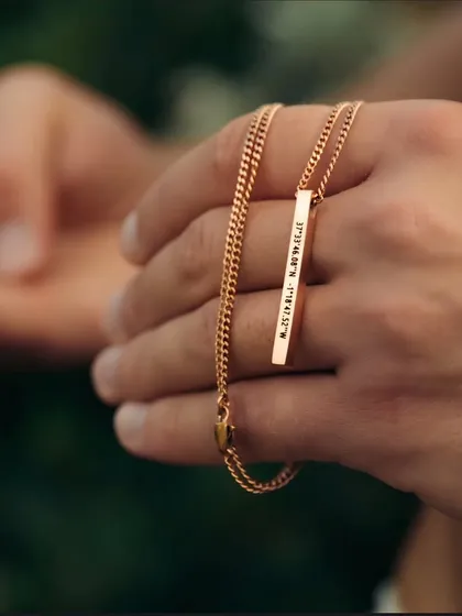 A hand holding a rose gold bar pendant engraved with coordinates. This shot highlights the warm tone of the rose gold finish and the precision of the custom engraving.