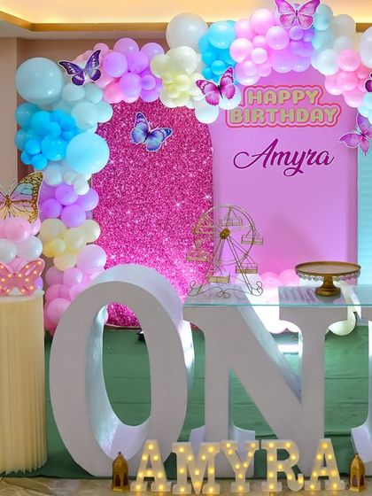 A beautiful butterfly-themed first birthday party. The decor features a stunning balloon arch in pastel colors, a sparkling pink backdrop, and elegant butterfly accents, creating a whimsical and enchanting atmosphere.