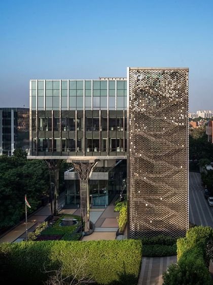 An elevated view of the Stainless Steel Office, highlighting the relationship between the main glass structure, the surrounding greenery, and the kinetic 'shimmer screen' on the fire staircase.