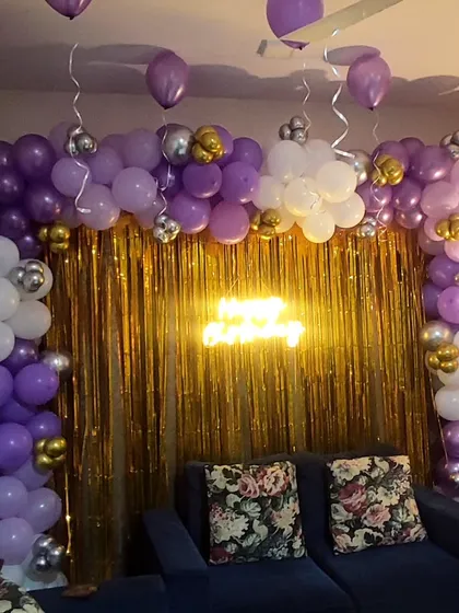 This birthday decoration uses a purple, white, and silver balloon garland over a gold shimmer curtain. A 'Happy Birthday' neon sign adds a modern and stylish focal point to the setup.