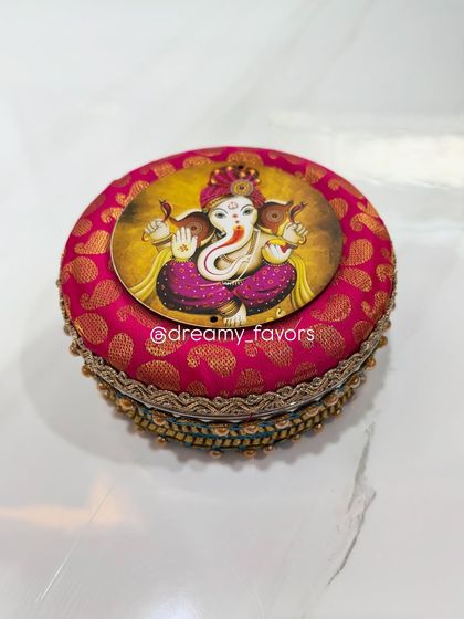 A detailed look at the lid of my handcrafted Modak box. It's covered in beautiful pink brocade fabric and features a Ganpati image on top, making it a special container for your festive sweets.