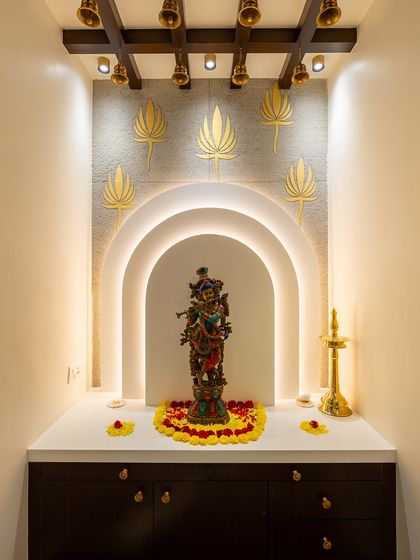 This pooja niche uses a beautiful archway with halo lighting to create a divine glow around the Krishna idol. Golden lotus motifs on the wall and traditional brass bells add to the sacred, traditional feel.