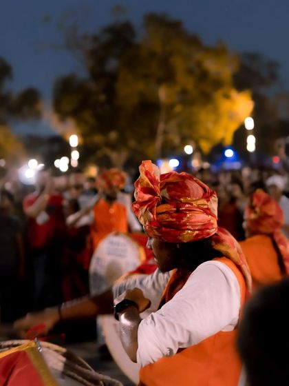 A dhol player is captured from the side, with the blurred motion of the crowd and trees in the background. This gives a sense of the lively, festive environment.