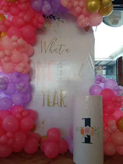 A "What a Wonderful Year" theme for a first birthday. This setup features a custom backdrop and a balloon arch in shades of pink, purple, and gold, with a small pedestal for the cake.