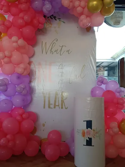 A lovely 'What a Wonderful Year' theme to celebrate a first birthday. This setup includes a custom backdrop, a pedestal for the cake, and a balloon garland in shades of pink, purple, and gold.