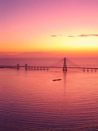 A breathtaking aerial view of the Bandra-Worli Sea Link in Mumbai, silhouetted against a fiery pink and orange sunset reflecting on the calm sea.