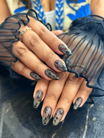 An elegant and sophisticated design featuring almond-shaped nails with a sheer, smoky gray base. Each nail is adorned with delicate, hand-painted black lace-like floral patterns, creating a timeless and ethereal look perfect for any occasion.