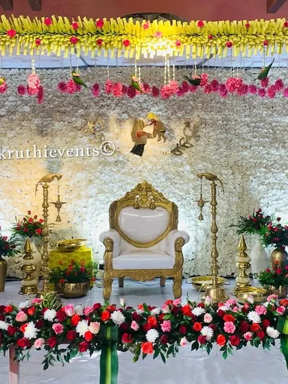 A classic stage for a traditional function, featuring a backdrop made of white flowers. The setup is framed with garlands of jasmine and roses, with a single ornate chair at the center.