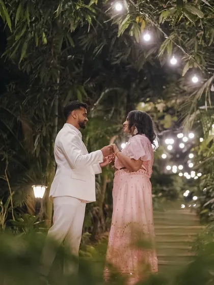 The couple shares an intimate moment in a lush, green pathway adorned with warm lights, creating a magical, fairytale-like setting.