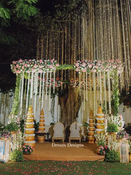 A full view of a traditional mandap setup under a canopy of twinkling lights. The design incorporates classic golden pillars and a profusion of pink and white flowers, creating a sacred and opulent space for the wedding ceremony.
