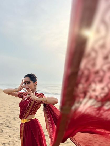 A candid moment during a beach photoshoot. The flowing saree against the sea breeze creates a beautiful, organic movement that complements the dance mudra.