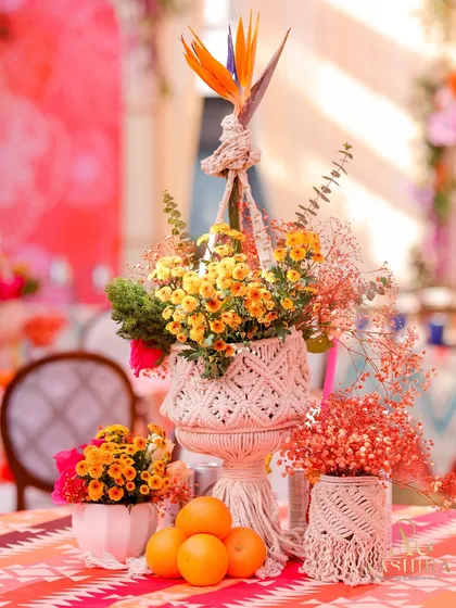 A whimsical centerpiece featuring a macrame plant hanger holding a pot of marigolds, surrounded by fresh oranges and other floral arrangements.