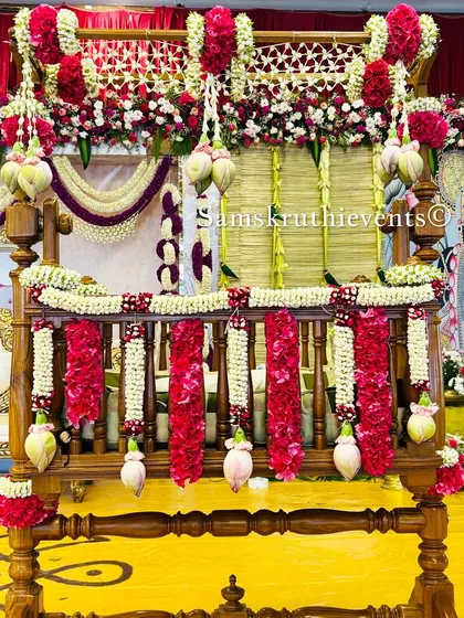 A close-up of a traditional wooden cradle decorated with red and white flowers. In the background, a Pichwai art panel and a yellow floor with rangoli add to the rich, cultural feel of the event.