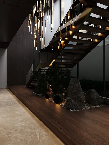 A zen-inspired indoor garden sits beneath a modern staircase. The dark wood, natural rocks, and dramatic chandelier create a moody, contemplative, and highly textural space.