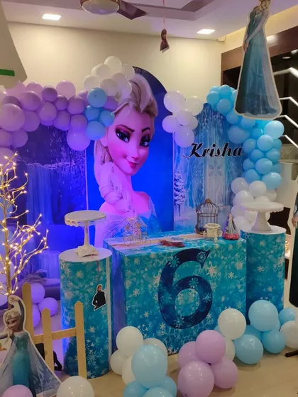 A detailed Frozen theme party with a large Elsa backdrop and glowing tree prop. The setup uses multiple pedestals and character cutouts to create a magical winter scene.