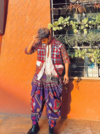 A cool, casual pose against a bright orange wall. This outfit features a mirror-work jacket over a simple white shirt and purple dhoti pants, a great way to style a modern Garba look.