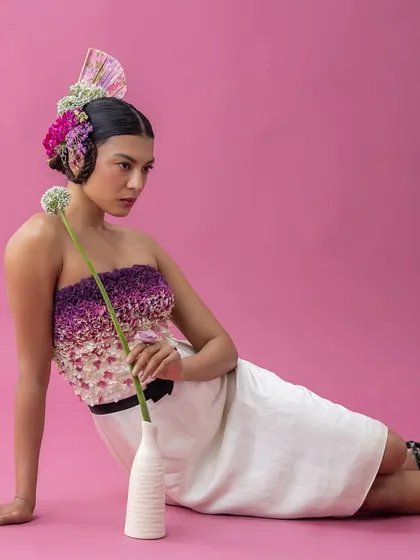 This posed shot from a high-fashion editorial combines a textured, strapless dress with a dramatic floral headpiece. The composition, with the model seated on the floor holding a single flower, was directed to create a feeling of grounded yet surreal elegance.
