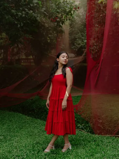 A serene portrait of Deyana in her vibrant red dress, standing against a lush, green backdrop. This image captures her quiet elegance and poise.
