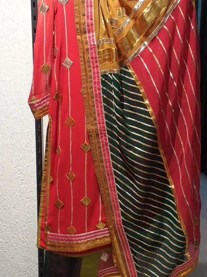 A vibrant Gota Patti suit by designer Preeti S Kapoor, featuring a red kurta and a multi-colored dupatta with green and red stripes. This is a perfect example of the renowned designer labels available in our outlet.