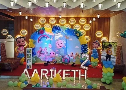 A full view of a Cocomelon stage decoration for a second birthday. The setup includes a large backdrop, balloon columns, character posters, and a light-up number '2' in a banquet hall setting.