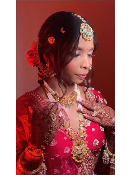 A profile shot capturing the intricate details of the hair and makeup. The soft curls and fresh flowers in her bun complement the timeless beauty of her red bridal attire.