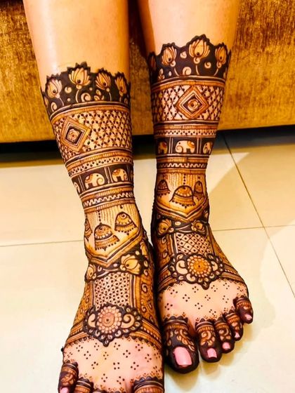 A close-up of beautifully decorated feet. The design features traditional motifs like bells (jhumkas) and elephants, with a neat border around the ankles.