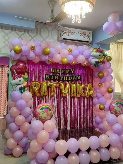 A unicorn-themed birthday decoration for a home party. It features a pink shimmer curtain backdrop and a balloon frame in pastel purple and pink.