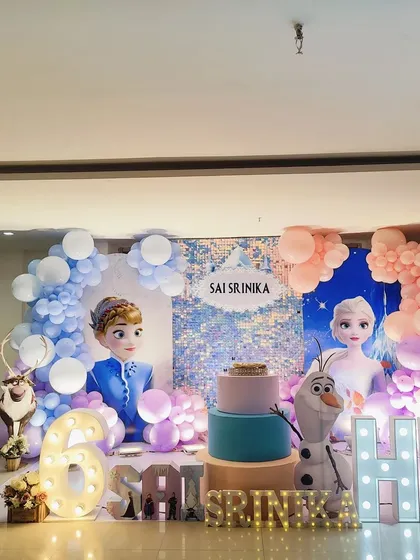 A magical Frozen birthday party setup with life-size cutouts of Elsa, Anna, Olaf, and Kristoff. The decor includes a shimmering sequin backdrop, themed balloon garlands, and light-up marquee letters.