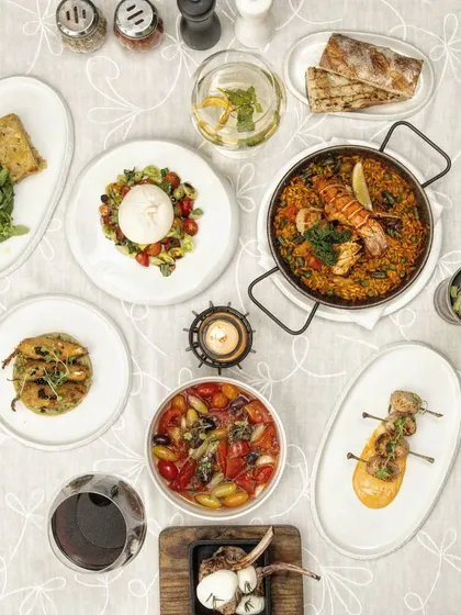 A beautiful flat lay of a Mediterranean feast, featuring paella, lamb chops, burrata, and various tapas. This image captures the variety and abundance of our menu.