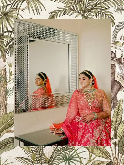 A moment of reflection for the bride. The ornate mirror frame complements her royal bridal look, featuring a heavily embroidered red lehenga and exquisite layered green bead jewelry.