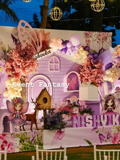 A magical fairy garden party for Nishvika, glowing under the evening sky. This enchanting setup includes a whimsical purple fairy house, large butterfly wing props, custom name lettering, and soft lighting from hanging lanterns to create a dreamlike atmosphere.