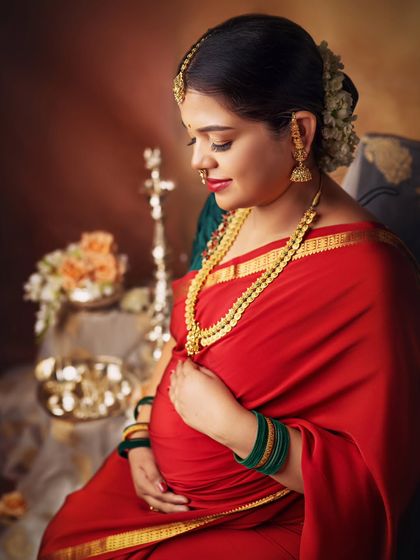 I love this pose, as the mom-to-be gently cradles her belly. The rich red saree and traditional gold jewelry beautifully complement her radiant glow in this classic maternity portrait.