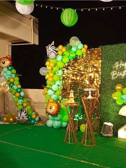 A wide view of an outdoor terrace jungle theme party for a second birthday. The setup includes a gold sequin and green wall backdrop, a light-up number '2', and a balloon garland that extends to a small teepee tent.