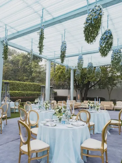 The guest seating area for the Anand Karaj, featuring elegant tables with blue linens and hanging floral installations overhead. The clear-top tent allows for an open, airy feel.