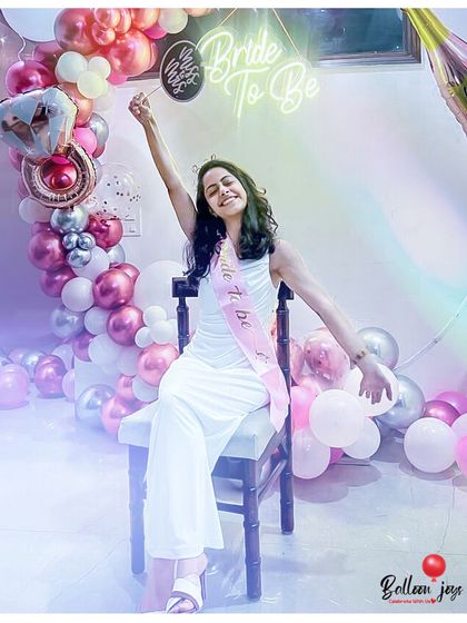 Blooming with good vibes before the big day! The bride-to-be is all smiles, sitting in front of a beautiful balloon arrangement with a 'Bride To Be' neon sign, ready to celebrate with her friends.