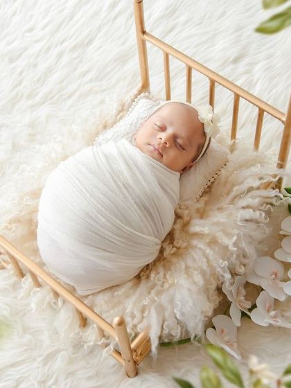 A newborn is swaddled in white and placed in a tiny golden cot, surrounded by soft white fur and delicate flowers for an angelic look.