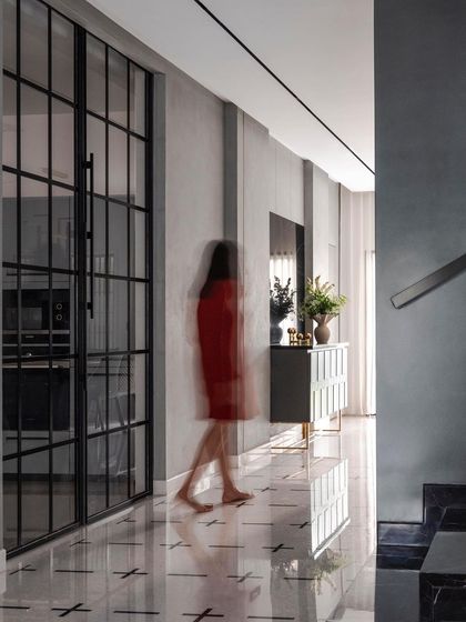 A long passage with polished marble floors and lime-plastered walls creates a central axis in the 'ARR' residence. Black-framed glass doors separate the kitchen while allowing light to pass through, maintaining a sense of openness and flow.