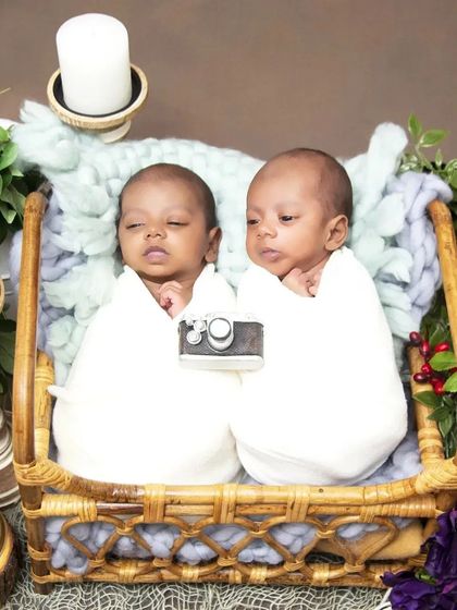 A simple and elegant setup for twin boys, both swaddled in clean white wraps. They are posed together in a basket, with one holding a tiny prop camera, perfect for a future photographer's first picture.