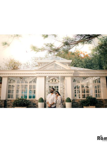 A wide shot of a couple standing before a beautiful white glasshouse, surrounded by trees. The bright, airy feel of this image is perfect for a romantic and elegant pre-wedding session.