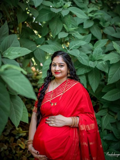 A close-up of the mother-to-be in her vibrant red saree, surrounded by lush green leaves. Her expression is one of serene confidence and joy.