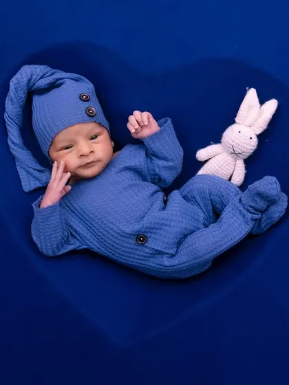 An alert newborn in a blue outfit lies in a heart-shaped prop, looking curiously at the camera.