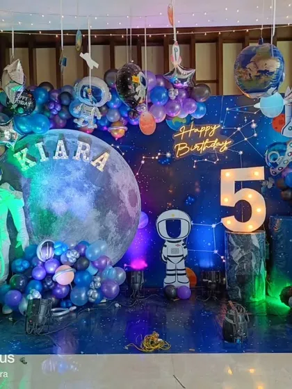 An out-of-this-world space theme for Kiara's 5th birthday. This decoration features a moon backdrop, astronaut cutouts, planet balloons, and a light-up number '5' for a cosmic celebration.