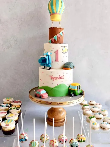 A fun vehicle-themed dessert table for a first birthday. The cake features a hot air balloon, scooter, and auto-rickshaw, with matching cupcakes, macarons, and cake pops.