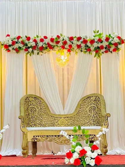 Here’s a more traditional and elegant stage I created, featuring a beautiful ornate sofa for the couple. The backdrop uses clean, golden rectangular frames accented with classic red and white flower arrangements to create a look of timeless grace for your special day.