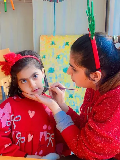 A teacher carefully paints a small Christmas tree on a student's cheek.